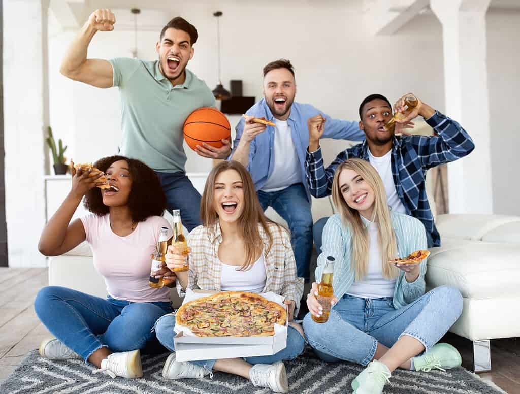 A group of six people enjoying pizza and drinks, with one person holding a basketball, while sitting and cheering in a living room setting.